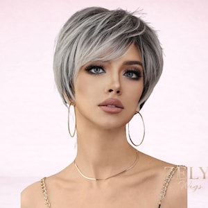 May include: A short, layered wig with a blend of gray and white tones. The wig features a side-swept bang and is styled to frame the face. The model is wearing gold hoop earrings and a gold necklace. The ZELYA Wigs logo is visible in the bottom right corner.
