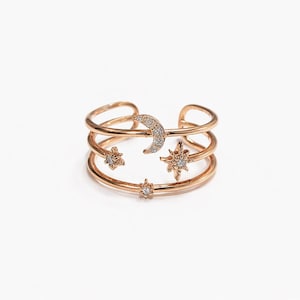 May include: Rose gold open ring featuring a crescent moon and star designs. The ring has three bands with a moon and three stars, each adorned with small, clear gemstones. The celestial-themed ring is a stylish accessory.