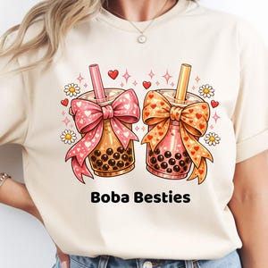 May include: A cream-colored t-shirt featuring a graphic of two boba tea drinks. Each drink is adorned with a large bow and a straw. The design includes hearts, daisies, and the text "Boba Besties" in black.