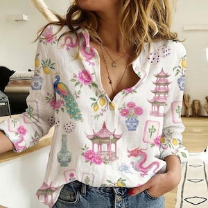May include: A white long-sleeve button-down shirt featuring a colourful print of pink pagodas, blue and yellow peacocks, dragons, floral vases, dice, and mahjong tiles. The shirt has a classic collar, a front button placket, and long sleeves.