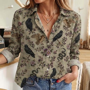 May include: A long-sleeved button-down shirt in a muted beige tone featuring a repeating pattern of black ravens, brown owls, winding snakes, and small purple flowers. The shirt has a classic collar, a front button placket, and cuffed sleeves.
