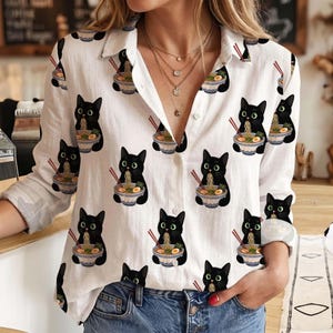 May include: A white long-sleeve button-down shirt featuring an all-over print of black cats eating bowls of ramen noodles with red chopsticks. The shirt has a classic collar, a front button placket, and long sleeves with buttoned cuffs.