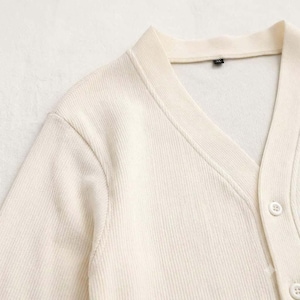 May include: A cream-colored, ribbed knit cardigan featuring a V-neckline and a button-down front. The garment includes a visible interior tag labeled XL, indicating the size. The fabric has a vertical texture throughout the sleeves and bodice.