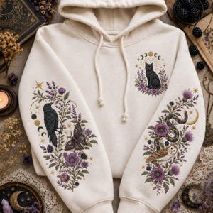 May include: A cream-colored hooded sweatshirt featuring intricate embroidery on the sleeves and chest. The designs include a black cat, a raven, a moth, a snake, and an owl, all surrounded by purple floral motifs, stars, and lunar phases.