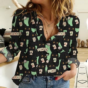 May include: A long-sleeved V-neck shirt featuring a repeating pattern of black cats on a light green background. Each cat is depicted holding various mahjong tiles, with some cats also shown with dice. The shirt has a button-down front placket.