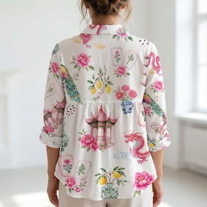 May include: A white long-sleeve button-down shirt featuring a colorful print of pink peonies, red dragons, pagodas, lemons, and mahjong tiles. The shirt has a collared neckline, a buttoned front, and long sleeves with rolled cuffs, showcasing a vibrant pattern.
