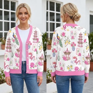 May include: A white V-neck cardigan with a pink trim and button closure, featuring a repeating print of pink pagodas, dragons, peacocks, mahjong tiles, and floral arrangements. The long-sleeved knit garment has a ribbed hem and cuffs.