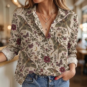 May include: A long-sleeved button-down shirt in a cream color featuring a repeating pattern of green luna moths, small purple butterflies, dark pink flowers, green foliage, and golden crescent moons. The shirt has a classic collar and white buttons.