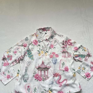 May include: A white long-sleeve button-down shirt featuring a repeating pattern of pink dragons, pagodas, blue and white vases, yellow lemons, peacock feathers, and mahjong tiles with Chinese characters printed across the fabric surface.