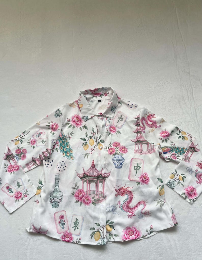 May include: A white long-sleeve button-down shirt featuring a repeating pattern of pink dragons, pagodas, blue and white vases, yellow lemons, peacock feathers, and mahjong tiles with Chinese characters printed across the fabric surface.