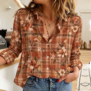 May include: A long-sleeved button-down shirt featuring a rust-orange and brown plaid pattern overlaid with a floral print of white and orange blossoms. The shirt has a classic collar, a front button placket, and is paired with blue denim jeans.