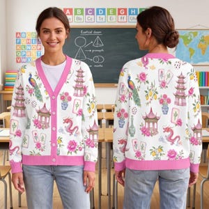 May include: A white long-sleeve cardigan with a pink V-neck trim and button closure. The fabric features a repeating pattern of pink peonies, pagodas, a red dragon, a peacock, lemons, and mahjong tiles against a white background.