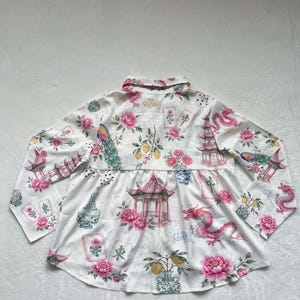 May include: A white long-sleeve button-down shirt featuring a repeating pattern of pink peonies, red dragons, pagodas, lemons, and mahjong tiles. The shirt has a collared neckline, long sleeves, and a gathered waist seam that creates a flared hemline.