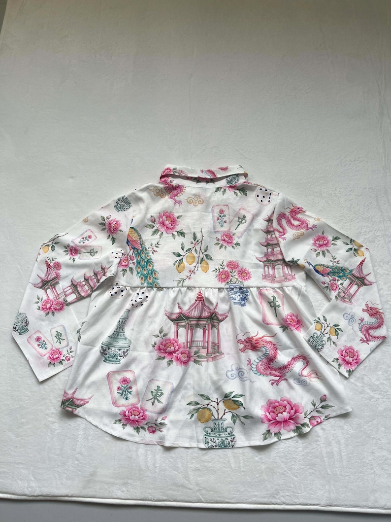 May include: A white long-sleeve button-down shirt featuring a repeating pattern of pink peonies, red dragons, pagodas, lemons, and mahjong tiles. The shirt has a collared neckline, long sleeves, and a gathered waist seam that creates a flared hemline.