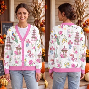 May include: A white V-neck cardigan with a pink trim and button closure, featuring a repeating pattern of pink pagodas, dragons, peacocks, mahjong tiles, floral vases, and lemons. The cardigan is worn over a plain white shirt and blue jeans.