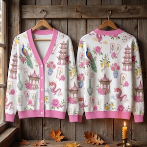 May include: A white cardigan with a pink V-neck and hem, featuring a repeating pattern of peacocks, dragons, pagodas, mahjong tiles, vases with flowers, and lemons. The cardigan has a button-down front with five white buttons and long sleeves.