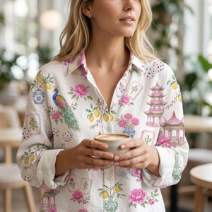 May include: A white long-sleeve button-down shirt featuring a colorful print of peacocks, pink flowers, yellow lemons in vases, pagodas, and mahjong tiles. The shirt has a classic collar and a buttoned front, showcasing a detailed pattern.