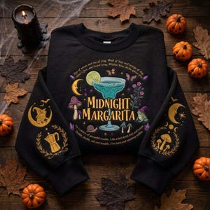 May include: A black long-sleeve sweatshirt featuring a graphic of a blue margarita glass, mushrooms, a crescent moon, and the text "Midnight Margarita." The sleeves are decorated with gold-colored prints of moons, mushrooms, and a coffee pot.