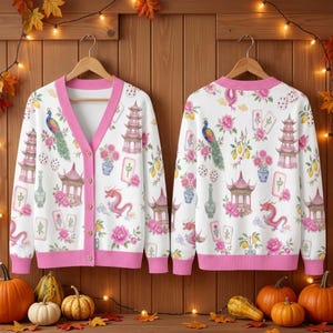 May include: A white V-neck cardigan with a pink trim and button front, featuring a repeating pattern of pink pagodas, peacocks, dragons, floral arrangements, and mahjong tiles. The cardigan is displayed on two wooden hangers side by side.