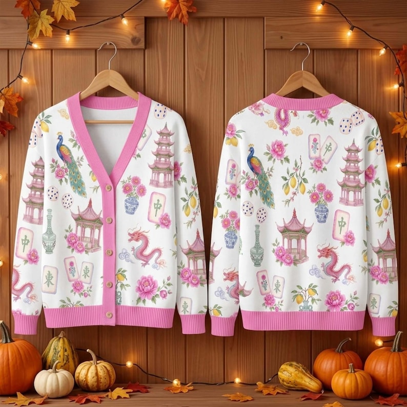 May include: A white V-neck cardigan with a pink trim and button front, featuring a repeating pattern of pink pagodas, peacocks, dragons, floral arrangements, and mahjong tiles. The cardigan is displayed on two wooden hangers side by side.