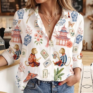 May include: A white long-sleeve button-down shirt featuring a colorful print of monkeys, pagodas, blue and white porcelain vases, cherry blossoms, and mahjong tiles. The shirt has a collar, a buttoned front, and cuffed sleeves.