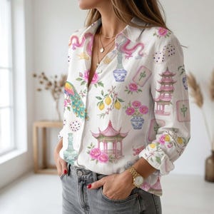 May include: A white long-sleeve button-down shirt featuring a colorful print of pink pagodas, blue and white vases, a peacock, lemons, mahjong tiles, dice, and pink flowers. The shirt has a classic collar and is paired with grey denim jeans.