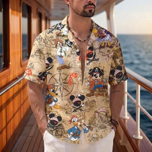May include: A short-sleeved button-down shirt in a tan color featuring an all-over print of cartoon characters dressed as pirates, treasure maps, compasses, skulls, and crossbones. The shirt has a classic collar and a front button closure.