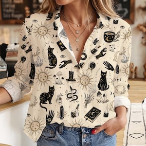 May include: A cream-coloured long-sleeve button-down shirt featuring a repeating black and gold print of celestial suns, moons, black cats, crystals, mushrooms, candles, books, hands, and potion bottles. The shirt has a classic collar and cuffs.