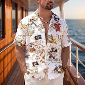 May include: A short-sleeved button-down shirt in white featuring a repeating print of cartoon characters, pirate ships, treasure maps, compasses, and black skull and crossbones flags. The shirt has a notched collar and a relaxed, open-front design.