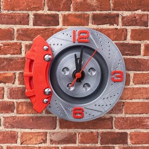 May include: A circular wall clock designed to resemble a silver automotive brake rotor with a red brake caliper attached to the left side. The clock face features red numerals at the 12, 3, and 6 o'clock positions, with black hour and minute hands.