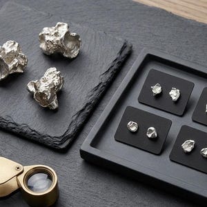Puede incluir: Piezas de joyería plateadas de forma irregular, incluyendo pendientes y objetos decorativos más grandes, se exhiben sobre una superficie gris oscuro. También hay una lupa con un marco dorado, lo que sugiere un enfoque en el detalle y la artesanía.