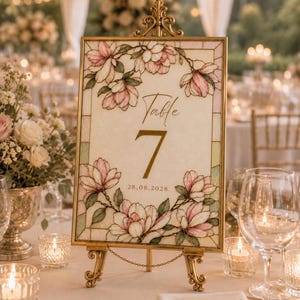Könnte beinhalten: Ein rechteckiges Tischnummernschild mit der Aufschrift Table 7 und dem Datum 28.08.2028, gerahmt in goldfarbenem Metall mit verziertem Oberteil. Das Schild zeigt eine Illustration im Buntglasstil mit rosa Magnolien und grünen Blättern.