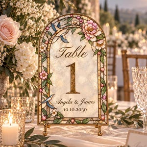 Könnte beinhalten: Ein bogenförmiges Tischnummernschild mit einem Rand im Buntglasstil, verziert mit rosa Blumen, grünen Blättern und blauen Vögeln. In der Mitte steht der Text Table 1, Angela & James, sowie das Datum 10.10.2030 in Schreib- und Serifenschrift.