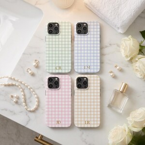 May include: Four phone cases with a gingham pattern in pastel colors: green, blue, pink, and beige. Each case has a gold monogram. Accessories include pearl jewelry, a perfume bottle, and white roses.