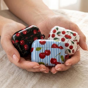 May include: Three AirPod cases with cherry designs. One case is black with red cherries, another is white with red cherries, and the third is blue and white striped with red cherries. The cases are held in open hands.
