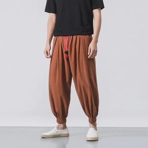 Op de afbeelding: Bruine harembroek met een rood trekkoord en zwarte kwast. De broek heeft elastische manchetten bij de enkels. De persoon op de afbeelding draagt een zwart t-shirt en witte schoenen. De broek is gemaakt van een zachte, soepele stof.