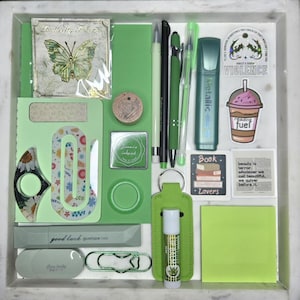 Puede incluir: Una colecci&oacute;n de art&iacute;culos de papeler&iacute;a verdes, que incluyen bol&iacute;grafos, un resaltador, pegatinas y un b&aacute;lsamo labial, dispuestos en una bandeja blanca. Los art&iacute;culos presentan textos como "Butterfly Flower", "good luck" y "reading fuel".