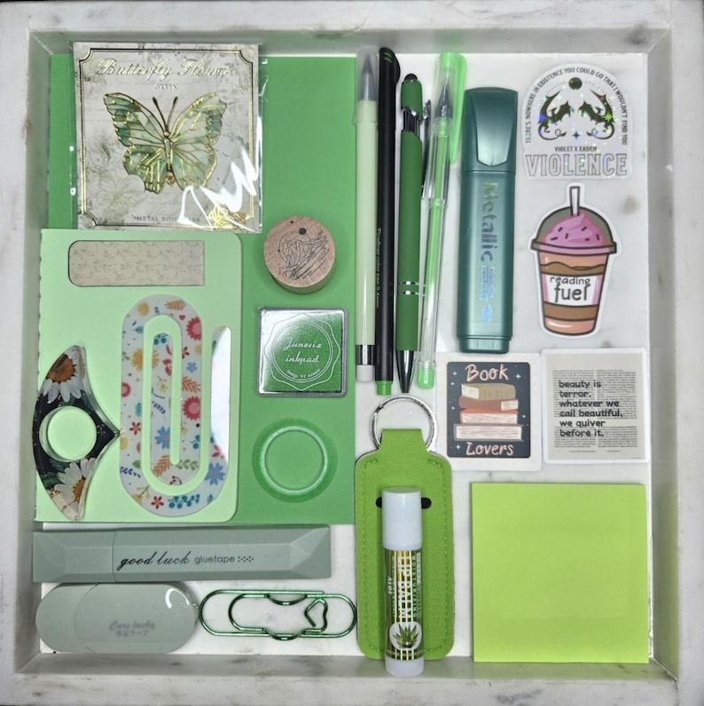 Puede incluir: Una colecci&oacute;n de art&iacute;culos de papeler&iacute;a verdes, que incluyen bol&iacute;grafos, un resaltador, pegatinas y un b&aacute;lsamo labial, dispuestos en una bandeja blanca. Los art&iacute;culos presentan textos como "Butterfly Flower", "good luck" y "reading fuel".