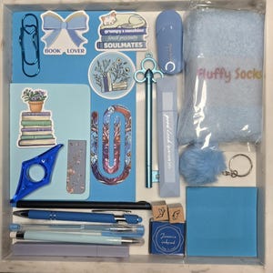 Puede incluir: Una colecci&oacute;n de art&iacute;culos de papeler&iacute;a y accesorios azules, que incluyen bol&iacute;grafos, un clip, pegatinas, un marcap&aacute;ginas, un resaltador, un llavero con pomp&oacute;n y calcetines esponjosos. Los art&iacute;culos est&aacute;n dispuestos en una bandeja blanca, con el texto "BOOK LOVER" y "Fluffy Socks".