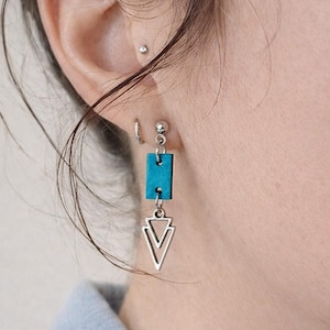 May include: A drop earring featuring a small, rectangular teal-coloured component attached to a silver-toned metal stud. Suspended from the teal rectangle is a silver-toned metal charm shaped like a double-lined triangle pointing downwards.