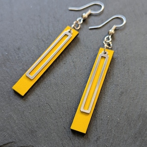 May include: A pair of drop earrings featuring long, rectangular yellow wooden pendants. Each pendant is accented with a smaller, centered silver-toned rectangular metal frame. The earrings hang from silver-toned metal fishhook ear wires.