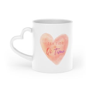 Könnte beinhalten: Weißer Keramikbecher mit herzförmigem Henkel. Der Becher hat ein Aquarell-Herz-Design in Pfirsich- und Rosatönen mit dem Text "Tea Time is Qi Time" in Rot und Lila.