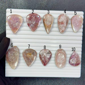 Pode incluir: Dez pedras preciosas polidas em forma de lágrima, de cor rosa e translúcida, dispostas em duas filas num expositor branco com ranhuras. Cada pedra está numerada de 1 a 10, exibindo inclusões únicas, tons variados de rosa e superfícies lisas.