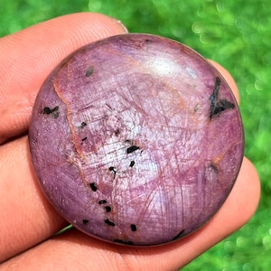 Könnte beinhalten: Ein runder, polierter Edelstein in tiefrosa Farbe, mit komplexen inneren Mustern und kleinen schwarzen Einschlüssen. Der Stein wird in einer Hand gehalten, wodurch seine glatte, reflektierende Oberfläche hervorgehoben wird. Der Edelstein ist ein Cabochon.