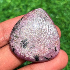 May include: A polished, teardrop-shaped gemstone with a swirling, textured surface. The stone is primarily a light purple color with silver and black inclusions. The gemstone is held in a hand against a blurred green background.