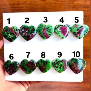 May include: Ten heart-shaped stones with a mix of green and red colors. Each stone is numbered from 1 to 10. The stones are arranged on a white surface, with a wooden background.
