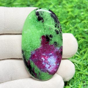 May include: An oval-shaped gemstone featuring a vibrant mix of colors. The stone has a base of bright green, speckled with black and deep red-purple hues. A patch of pale blue-white adds contrast. The polished surface reflects light, highlighting the natural patterns.