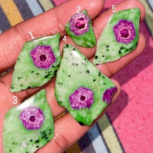 May include: Five polished, irregularly shaped cabochons of green zoisite with black speckles, each featuring a central hexagonal magenta ruby inclusion. The stones are numbered one through five in white text, displaying a smooth, glossy surface.