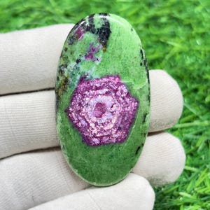 May include: Oval-shaped gemstone with a vibrant green base, speckled with black and featuring a prominent, textured, magenta-colored center. The gemstone is held in a white gloved hand, showcasing its polished surface and natural beauty.