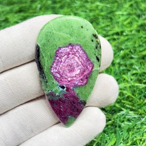 May include: A polished gemstone with a teardrop shape. The stone features a vibrant green base with a central, flower-like pattern in shades of pink and red. Black inclusions are scattered across the surface. The gemstone is held in a white glove.